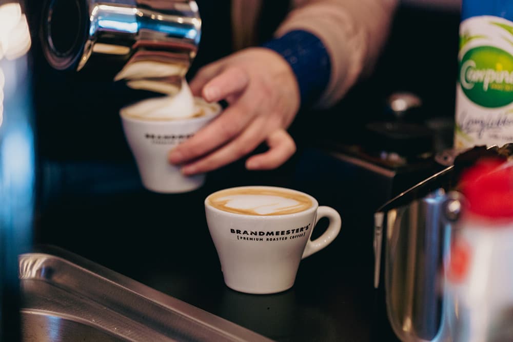 Melkschuim wordt in een kopje geschonken naast een cappuccino in een kopje van Brandmeesters