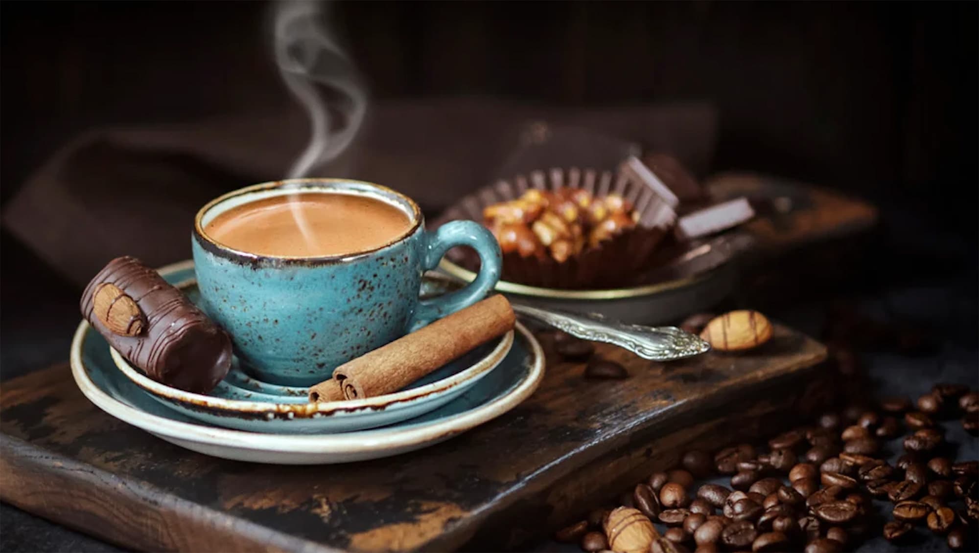 Een dampende kop koffie op een schoteltje met een kaneelstokje, omringd door koffiebonen en chocolade